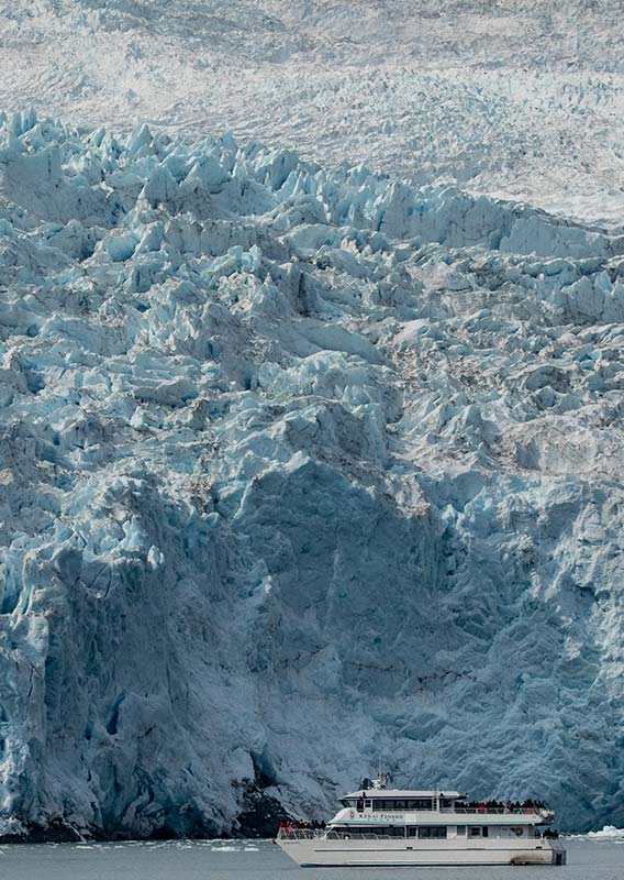 A boat below a massive glacier at the sea edge.