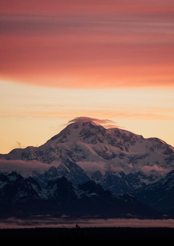 Denali, a large mountain rises high above clouds and below a red sky.