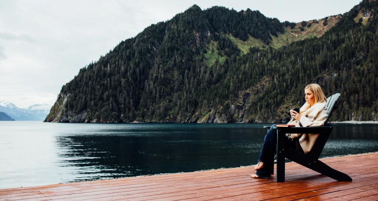 A woman sits in an Adirondack chair on a dock, looking out to ocean and tree-covered cliffsides.