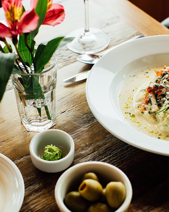 A dinner plate and olives on a dining table with a small vase of flowers.