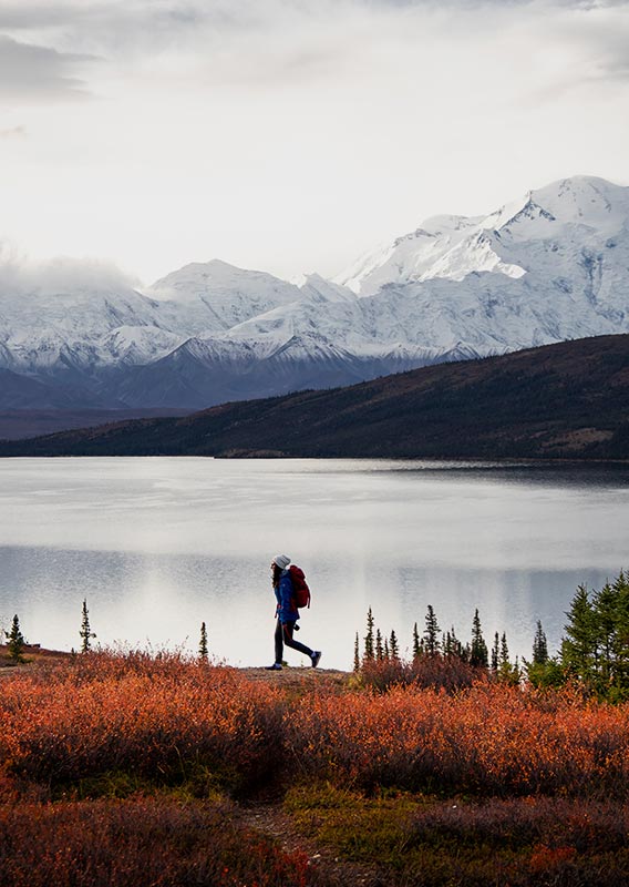 A person walks next to a lake below tall snow-covered mountains.