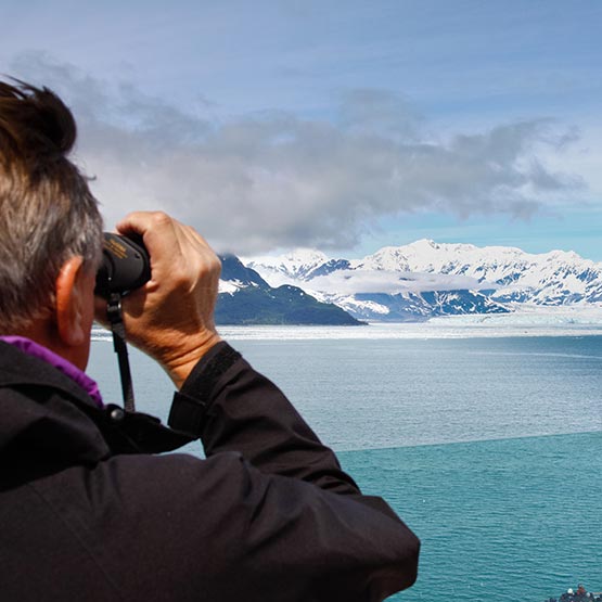 A person looks with binoculars at a glacier from a boat deck.