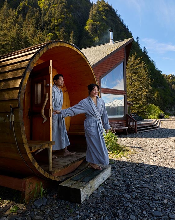 Two people exit a sauna onto rocky beach.